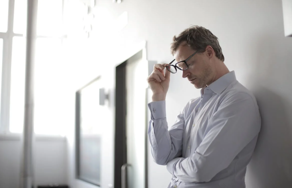 A main holding his glasses, leaning on a wall